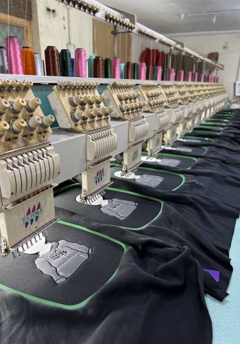 A row of industrial embroidery machines stitching intricate designs on black garments, highlighted by vibrant spools of thread in various colors in the background.