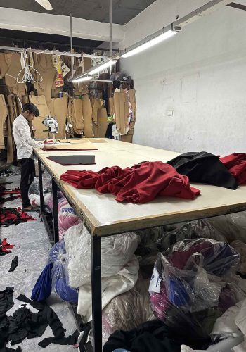 A worker in a garment factory focusing on cutting red fabric on a large table, surrounded by piles of fabric and patterns in a busy workshop.