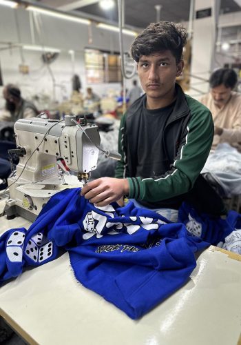 "A young male worker operating a sewing machine, focusing on a blue sports jersey adorned with dice and letters, in a busy garment factory.